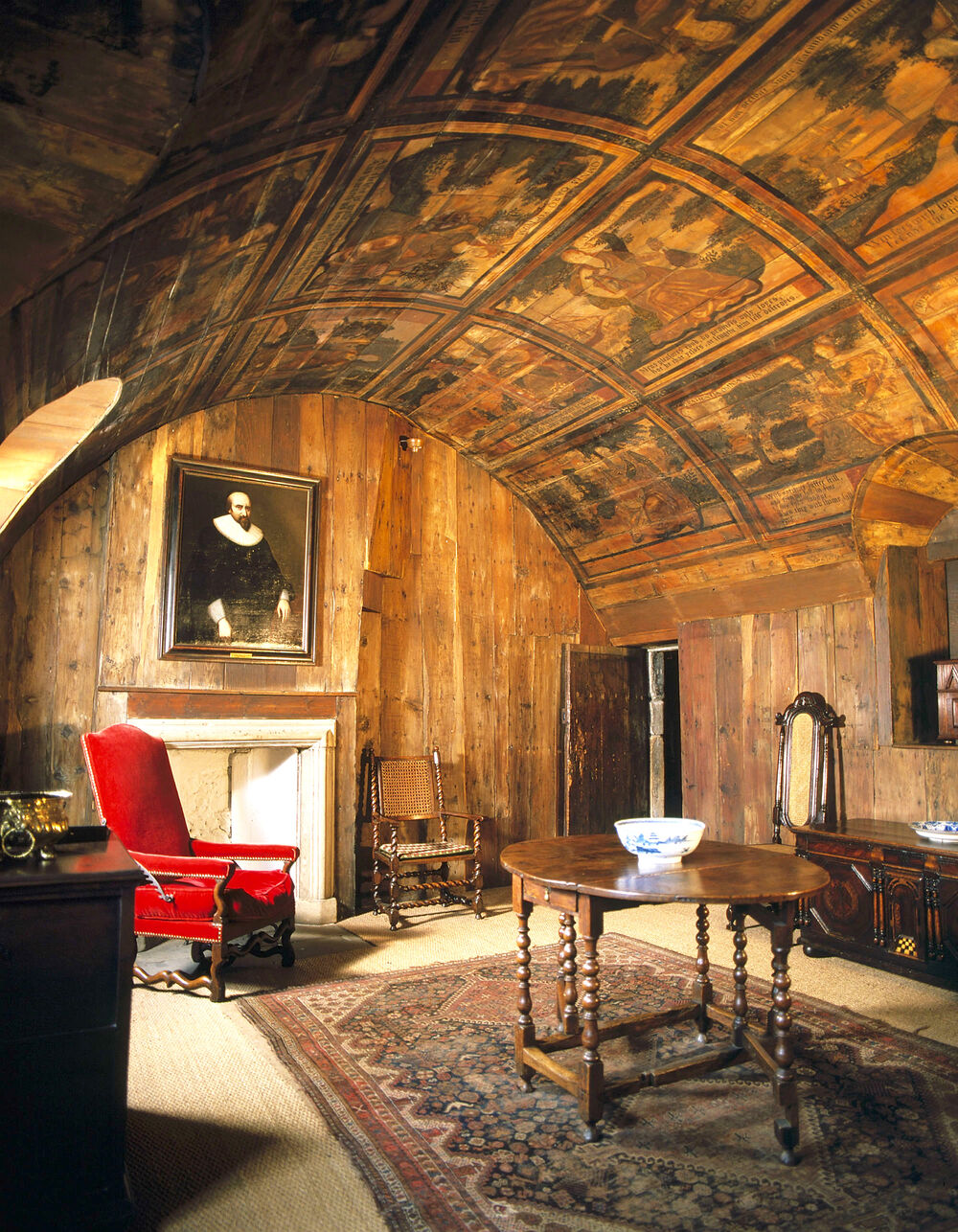 A room with a barrel ceiling and wood-panelled walls. The ceiling is covered in painted panels. A red armchair stands at the far end of the room. A wooden table stands in the centre of the room on a Turkish rug.