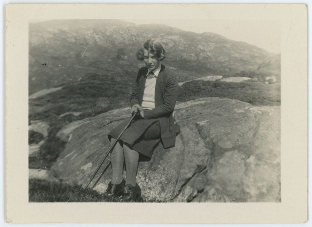 A black and white photograph of a young woman seated on a rock in a landscape. The woman has short bobbed hair and wears a dark jacket and skirt and a pale shirt. She holds a walking stick or cane in her lap with both hands.