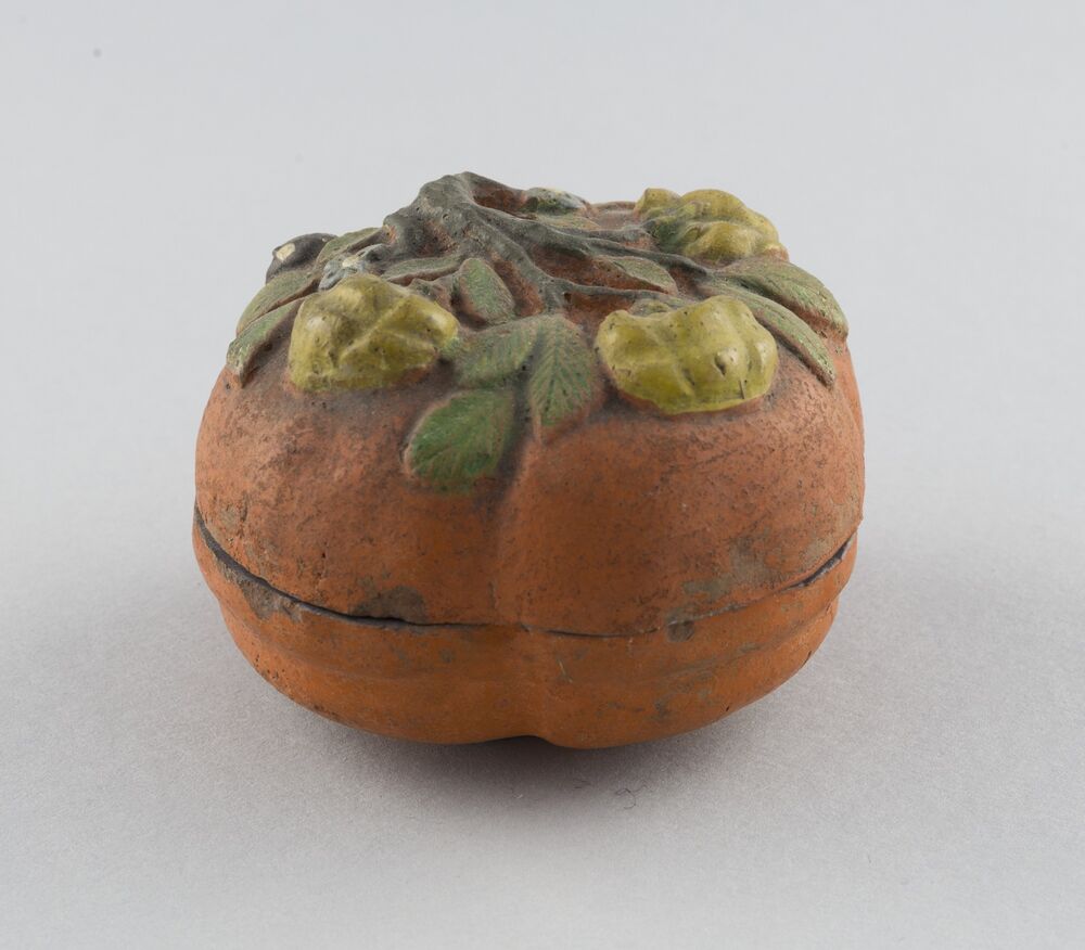 A ceramic pot in the shape of a squashed sphere, flat on the top and bottom. The pot is glazed in a dull, dusty orange and has a design on top of yellow flowers and green, oval shaped leaves.