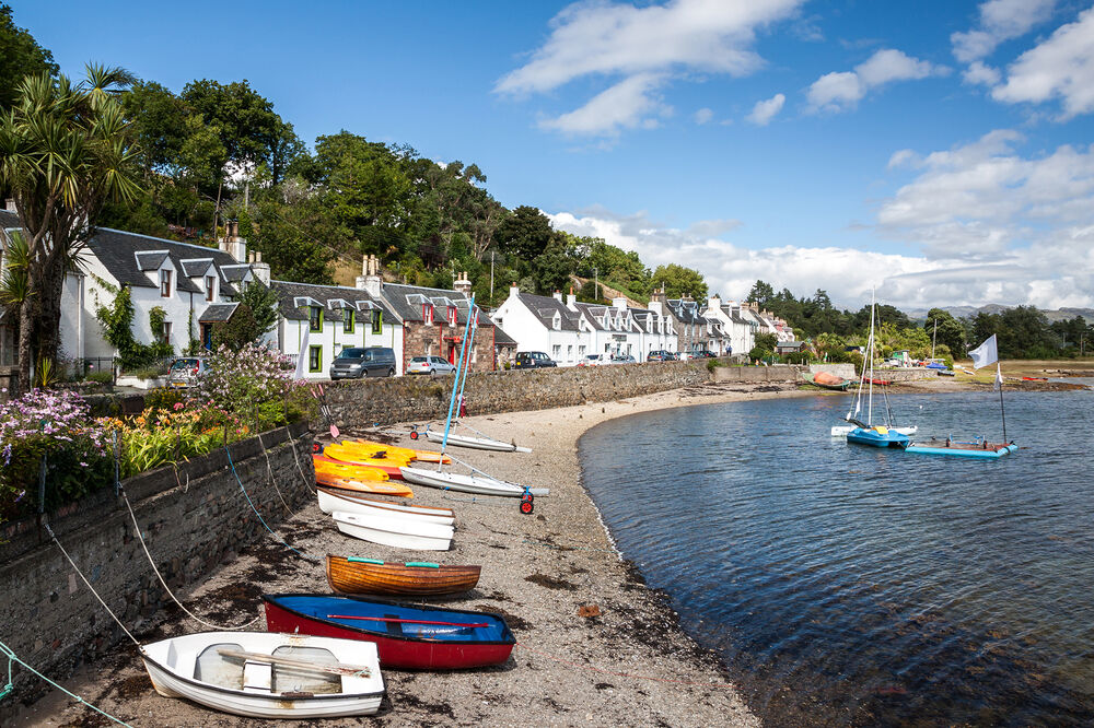 Wooden rowing boats are hauled up on the sandy beach in the bay in front of Plockton. The street faces the harbour and is lined with white cottages, with woodland rising behind.