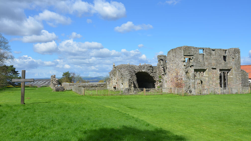 An old ruined abbey stands behind a grassy field on a sunny day. The ruins are encircled by a low wooden fence. There is a large wooden cross in the field in the foreground.