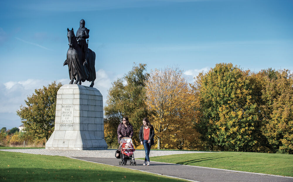 Two women, one with a pushchair, walk past the towering statue of Robert the Bruce atop his horse, on the hill at Bannockburn.