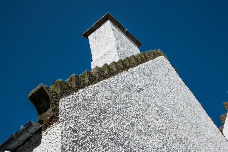 A detail of the roof of Abertarff House, shown against a bright blue sky. There is a dark crow-stepped gable line, between the white harled walls and chimney.