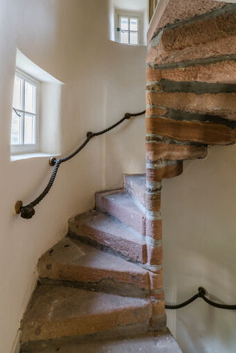 A steep stone spiral stonecase runs up a turret in an old house. A rope handrail runs along the outside wall.