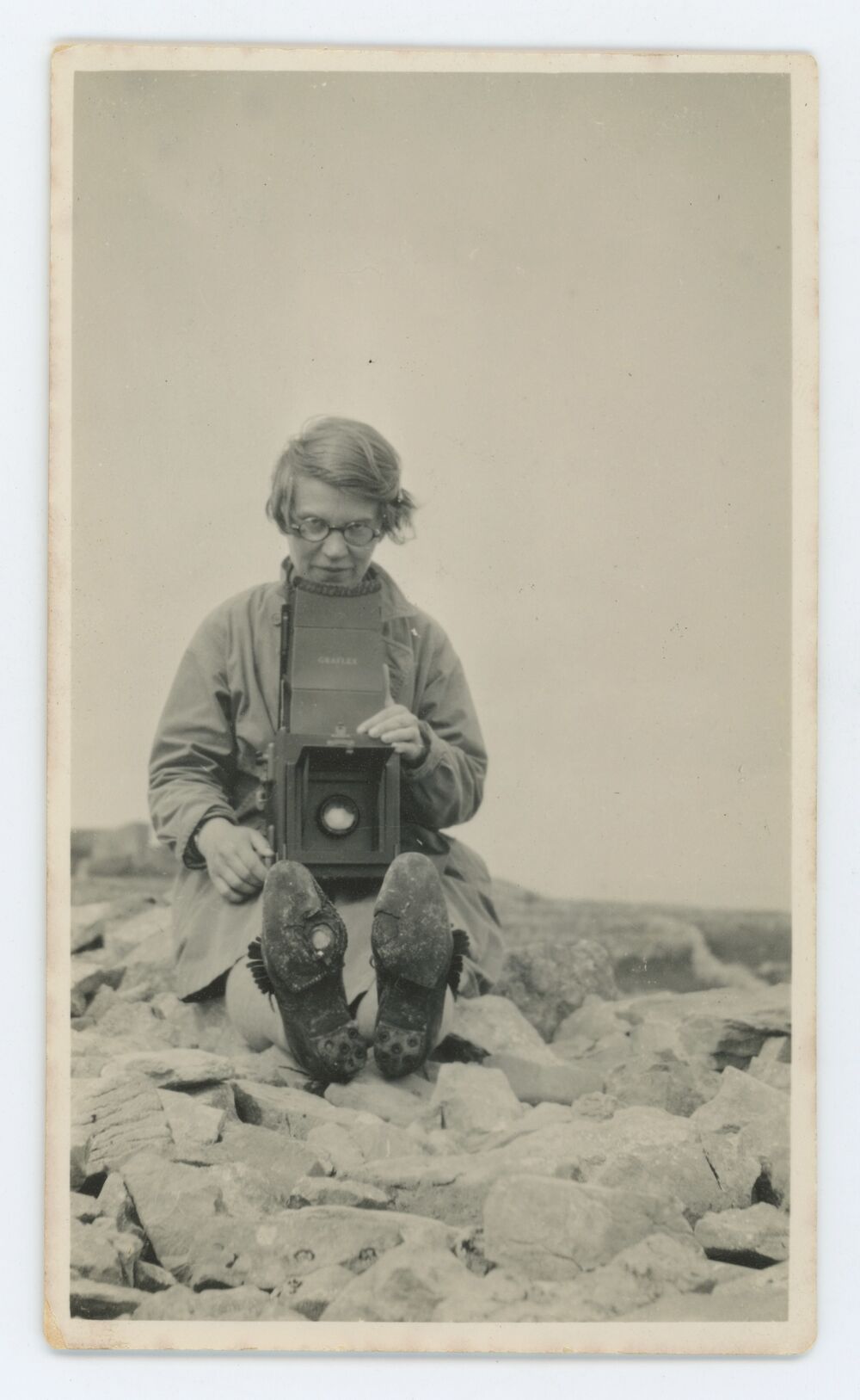 A sepia photo of a woman sitting on rocky ground. She holds a large camera and the soles of her boots face the viewer.