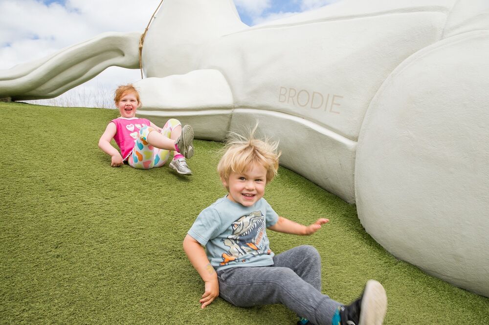 Two young children slide down a grassy slope beside a giant model of a white rabbit, that is lying on the bank.