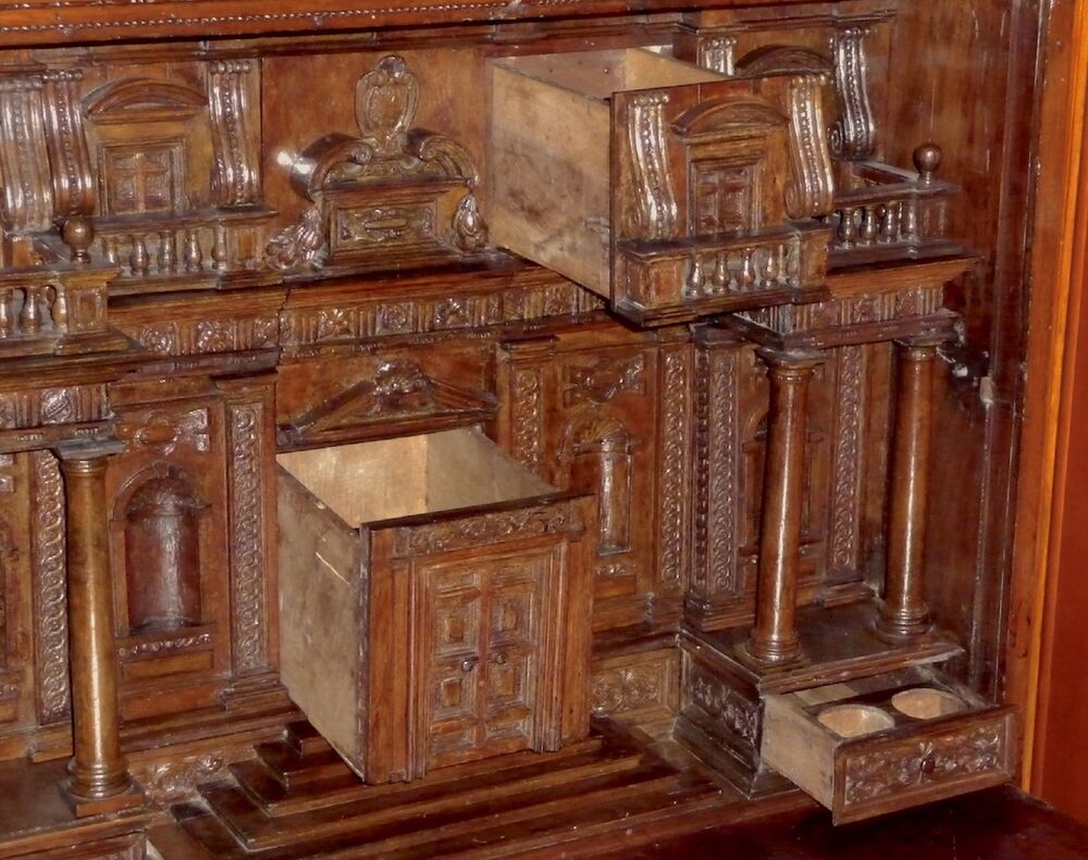 A close-up view of a wooden cabinet, opened up to reveal a collection of different shaped drawers. When closed, the drawers form the shape of a grand house with carved pillars. Two deep drawers in the middle are pulled open, as well as a small drawer on the bottom right, with two inkwell holes inside.