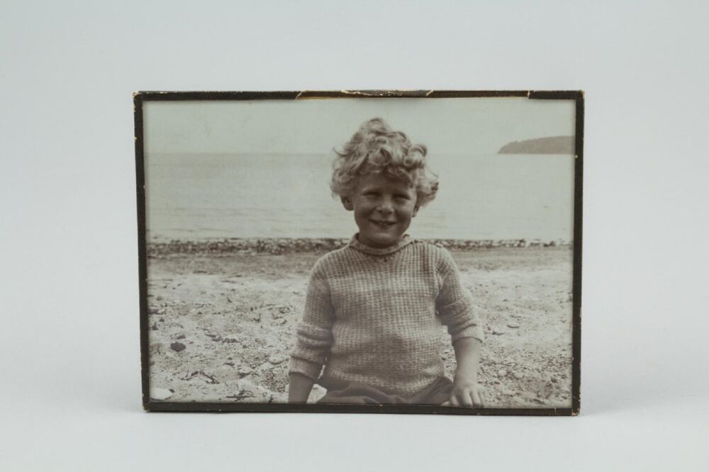 A black and white photograph of a young child, with curly fair hair, standing on a sandy beach. The photo is framed by a narrow black wooden frame, and is displayed against a plain grey background.
