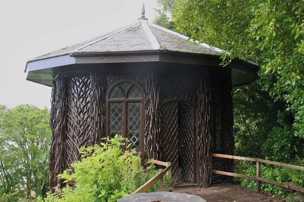 A wooden summerhouse building, with 10 sides, is perched at the edge of a rocky crag, with woodland behind. It has a conical roof. A wooden fenced path leads up to the door. The walls are intricately carved to depict vines and other creeping plants.