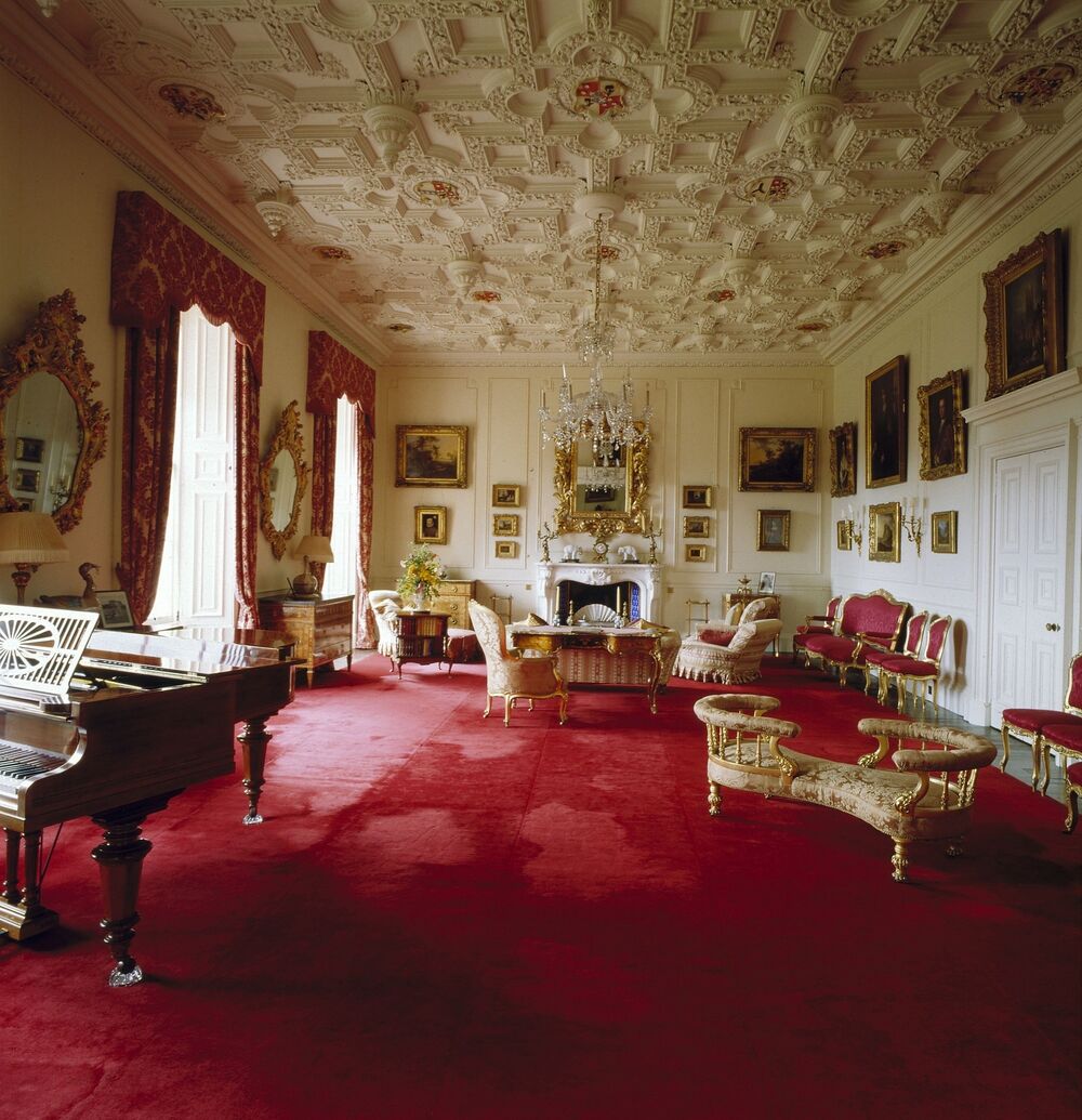 A view of a grand drawing room in Brodick Castle. It has a rich red carpet running the full length of the room. The plaster ceiling is intricately carved in panels with heraldic devices. A piano stands in the foreground, and further back are several sofas and chaise longues. The large windows are draped with long red curtains.