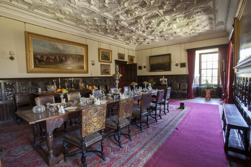 A view of the grand dining room in Brodick Castle. A long wooden dining table runs down the centre of the room, with glassware and napkins set upon it. The highly decorative plaster ceiling can be seen above. The lower half of the walls are covered with a dark wooden panelling, also with intricate carvings.