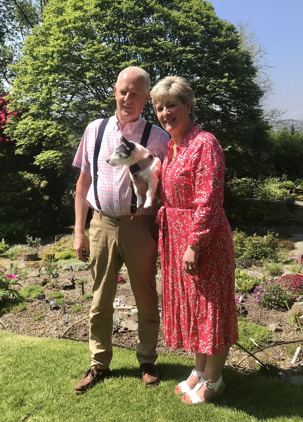 A man and a lady stand beside a rock garden area in Branklyn Garden on a sunny day. They carry a white and black Jack Russell terrier between them.