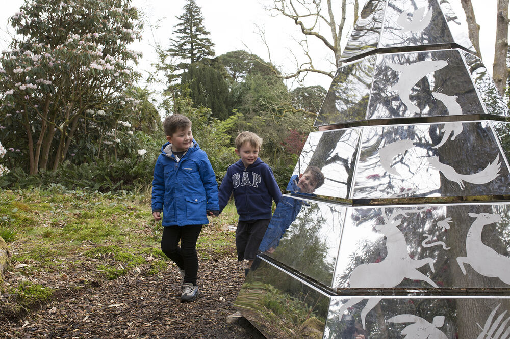 Two young boys walk hand in hand along a woodland path. They are approaching a mirrored glass pyramid sculpture.