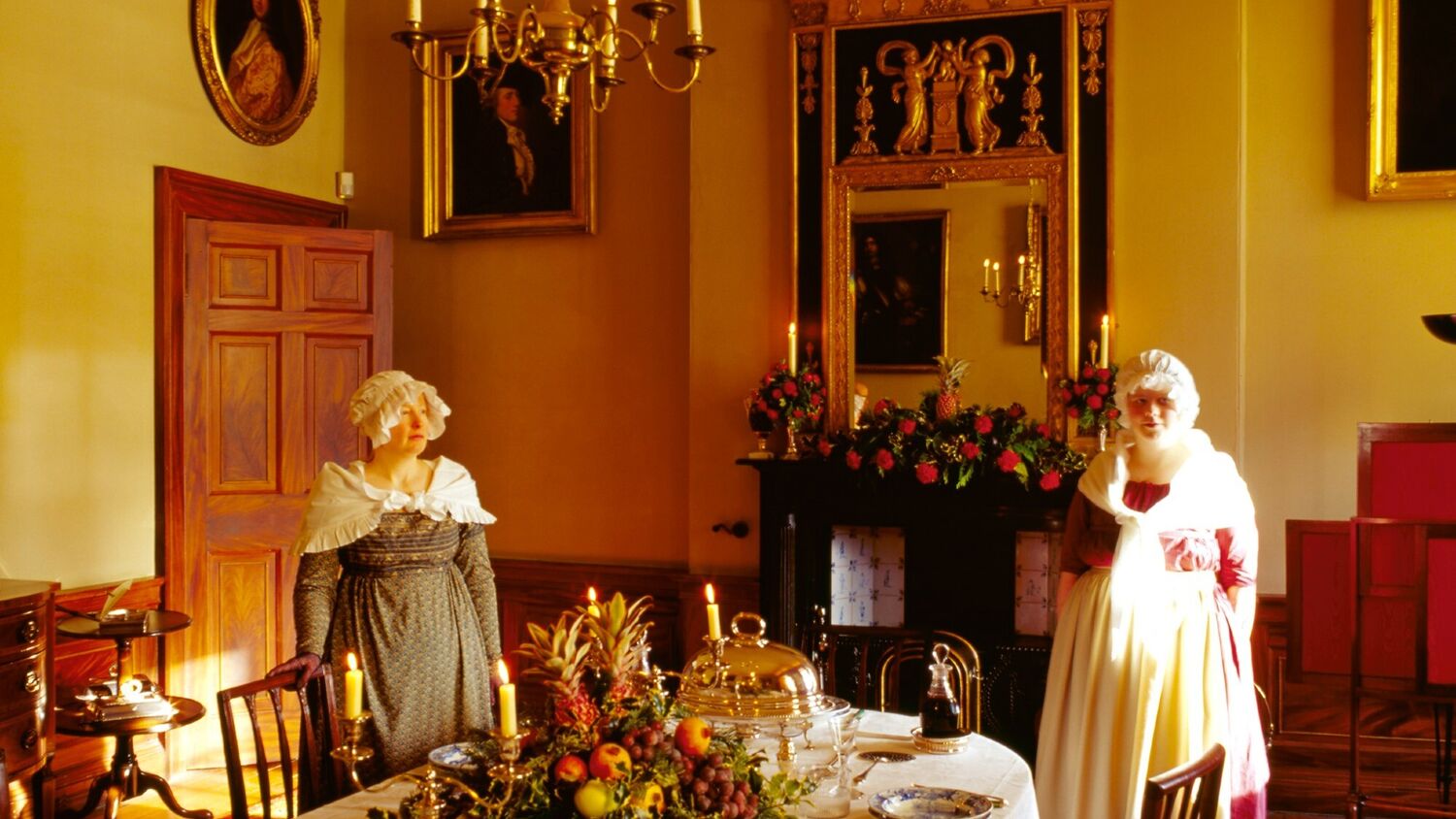 Two women in Georgian servants' costumes stand by a laid table in a grand Georgian dining room.