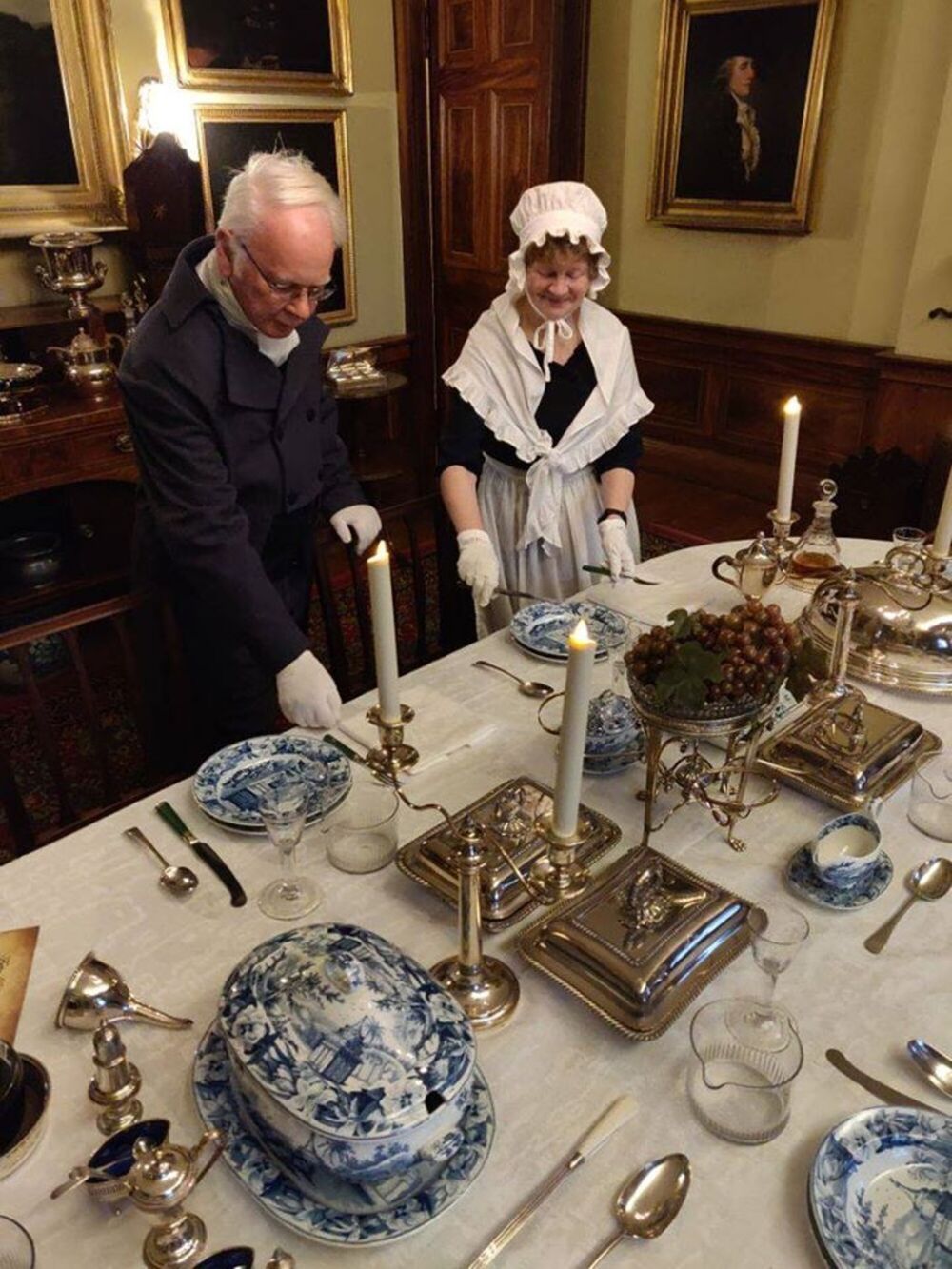 A butler and a housekeeper in Georgian costumes are laying a table for dinner in a grand Georgian dining room.