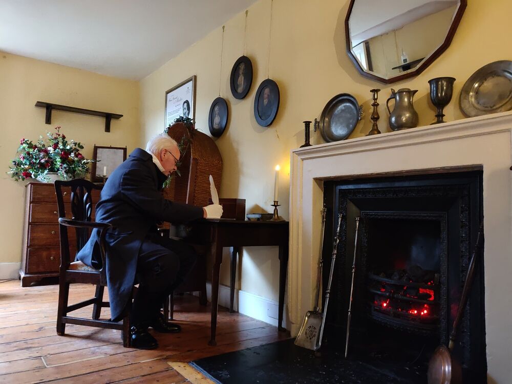 A man in a Georgian butler's costume is sitting at a table in a room with an open fireplace.