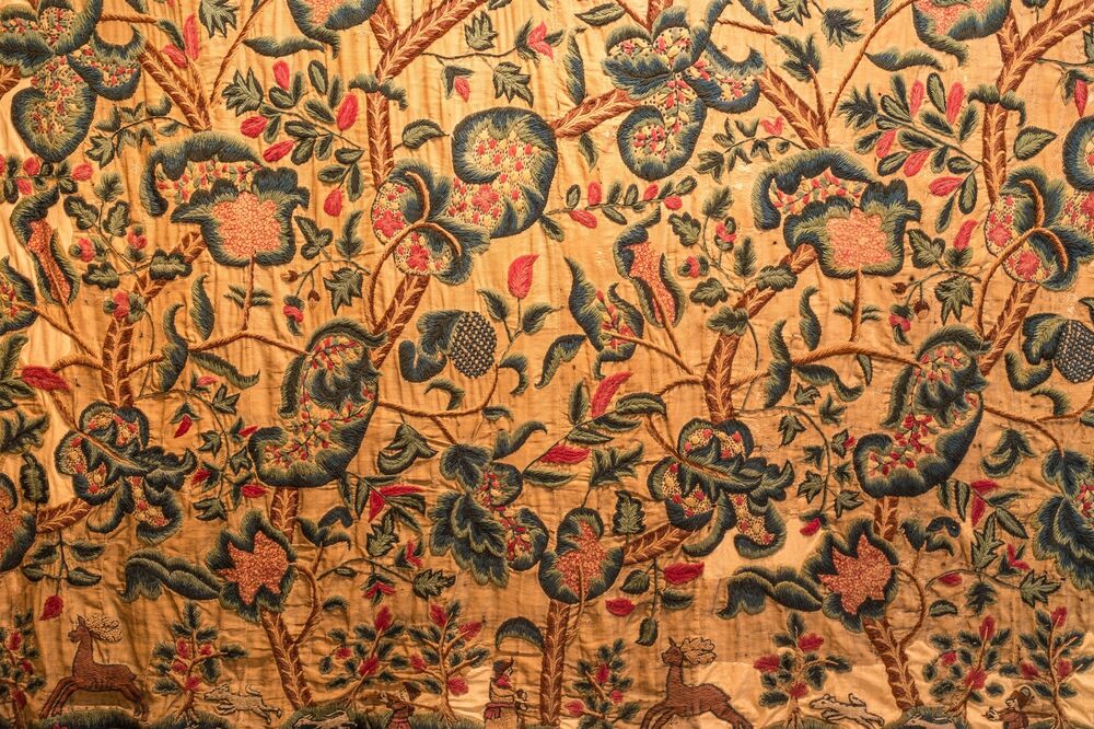 A close-up view of a tapestry panel, stitched in reds, greens and blues on a dark cream panel. There are lots of flower and leaf motifs, with some small deer, dogs and human figures at the very bottom.