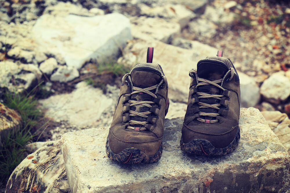 A pair of hiking boots sit side by side on a rock at the edge of a mountain path.
