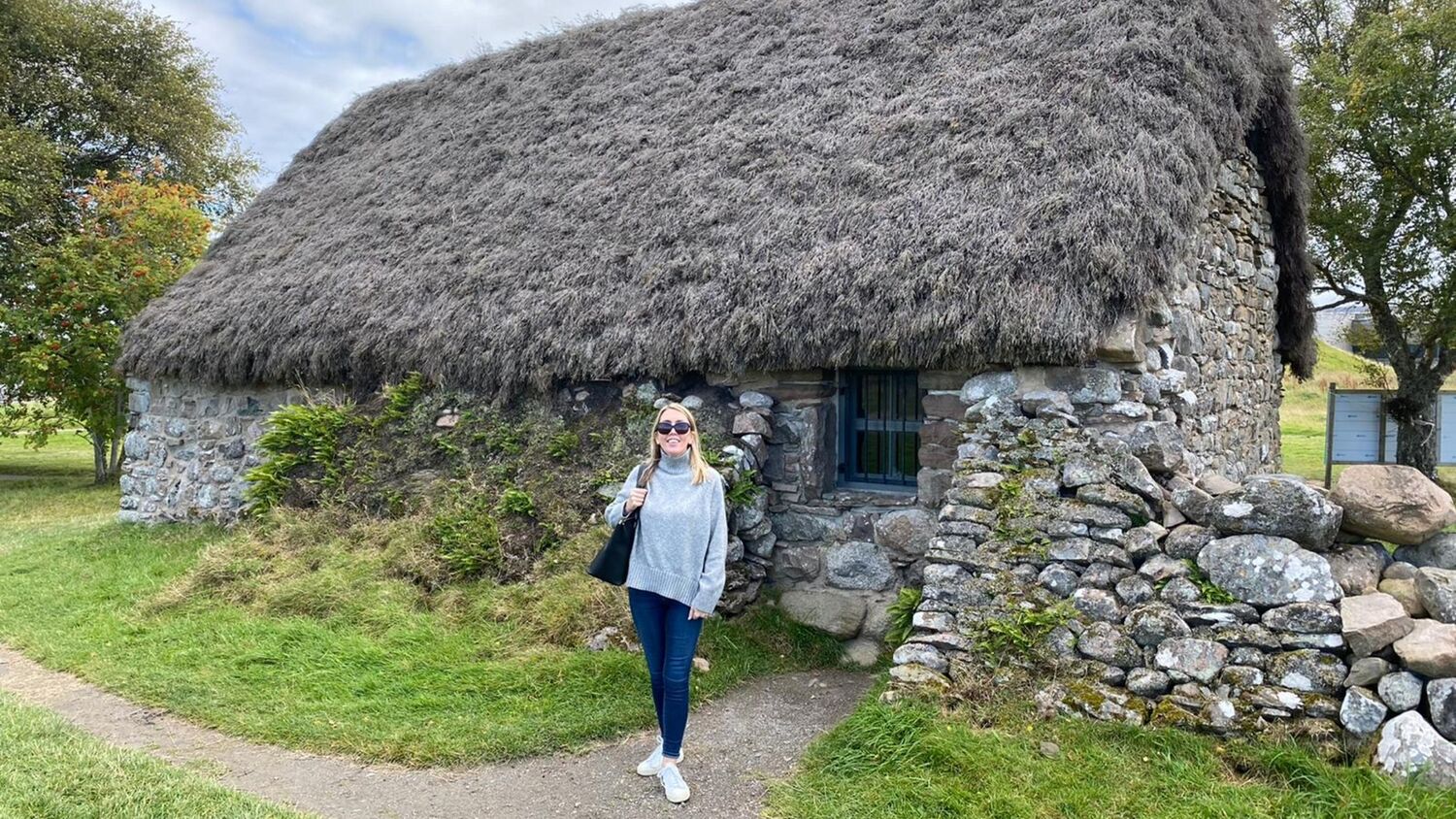 A smiling young woman stands on a narrow path outside a thatched cottage. It is a sunny day and she wears sunglasses.