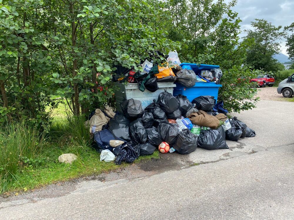 A large blue rubbish bin at a roadside overflowing with black rubbish sacks and other rubbish.