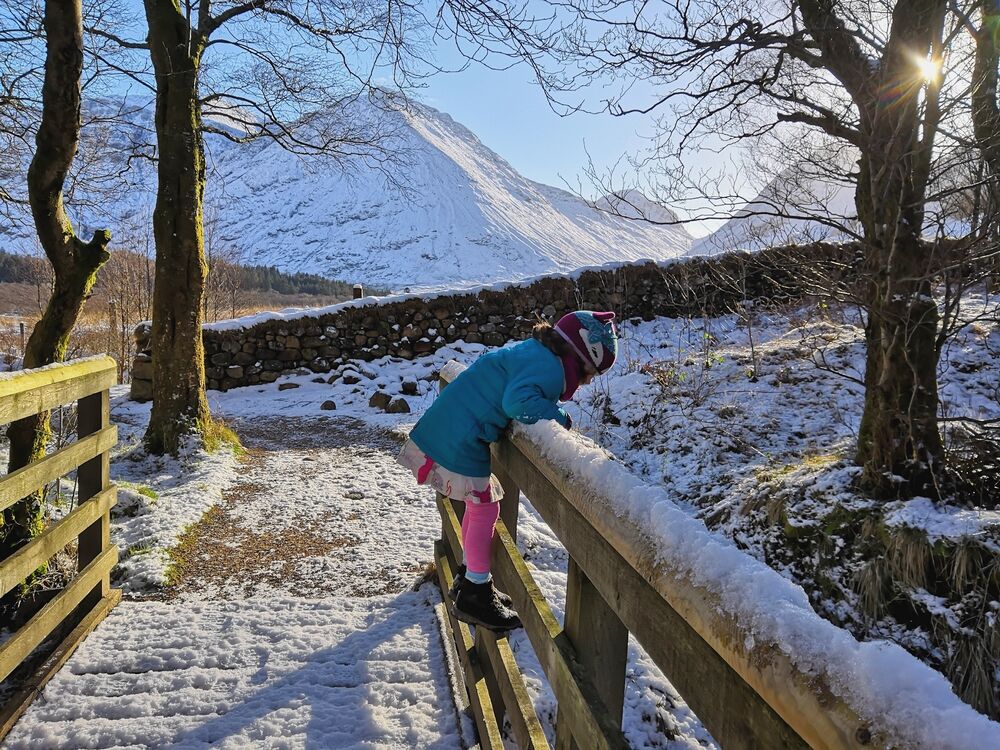 A little girl stands on a wooden bridge, looking over the railing below. The bridge and ground is covered in snow, and the tall mountains of Glencoe rise behind, forming a brilliant contrast against the bright blue sky.