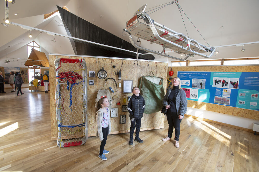 A mother and two children look up at a rescue sledge suspended from the ceiling in the new Glencoe Visitor Centre. It is an airy space with polished wooden floors and spaced out exhibits and panels.