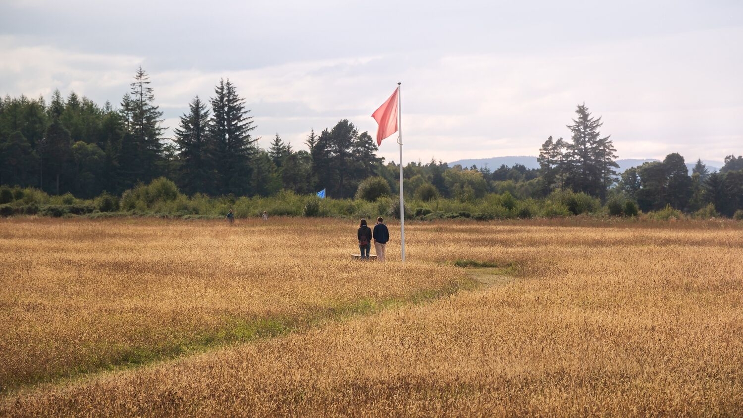 A couple stand on Culloden Battlefield beneath a tall flagpole from which flies a red flag. Woodland can be seen at the side of the field.