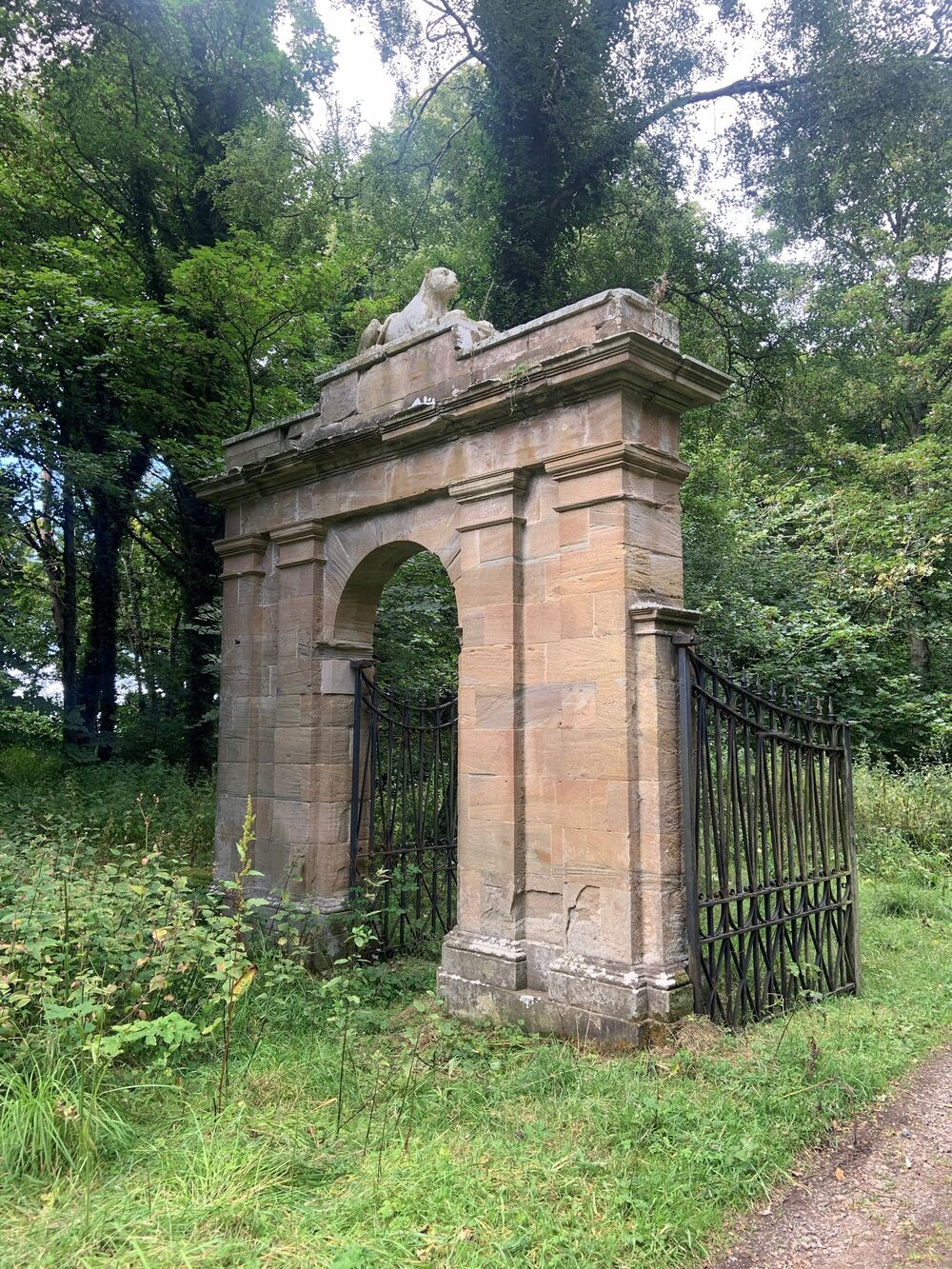An old stone gateway stands next to a track, with black wrought iron gates standing open. On top of the stone arch lies a stone statue of a large cat. Woodland is behind the track.