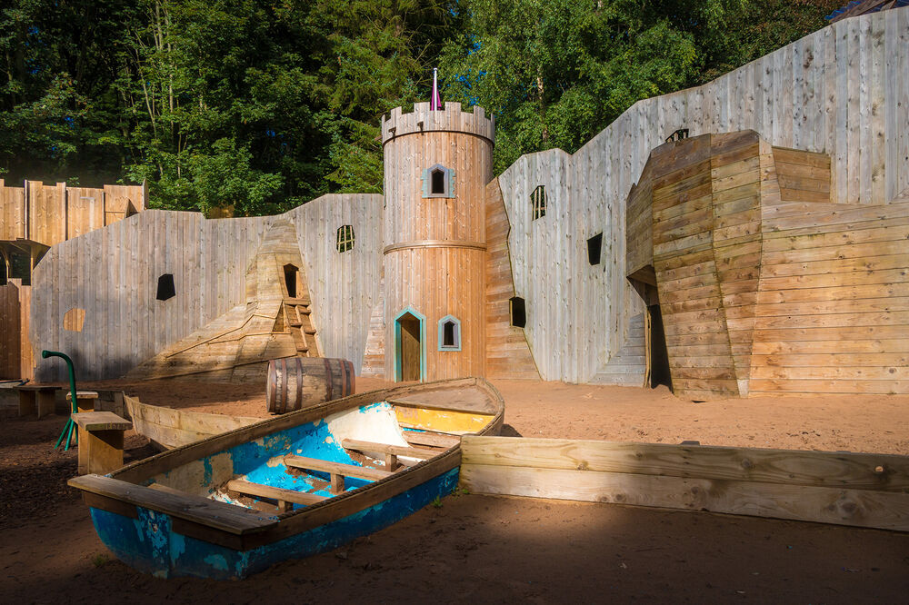 A rowing boat sits on a bed of golden sand. Behind it is a mini castle tower and a long wall made from wood.
