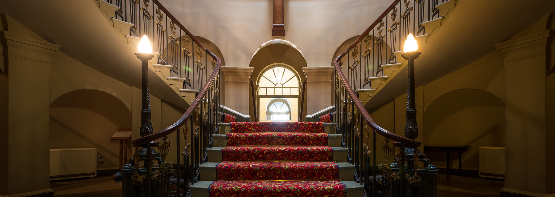 A view of the Oval Staircase at Culzean Castle, looking up from the ground floor. The wide sweeping stairs are covered by a red patterned carpet in the middle of the tread. Two standard lamps stand either side of the staircase.