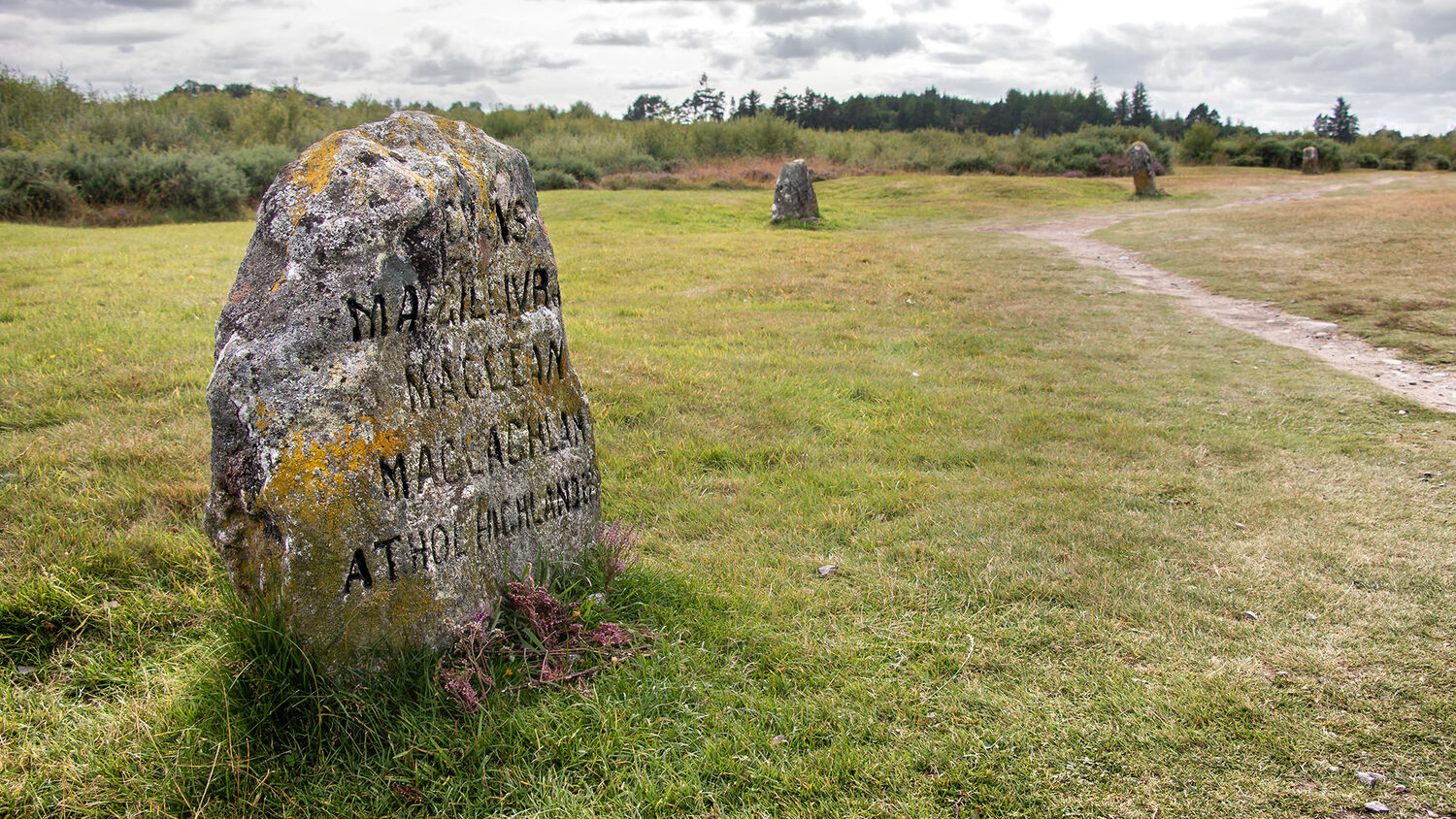 A gravestone stands beside a path on Culloden moor. The stone has lichen growing on it.