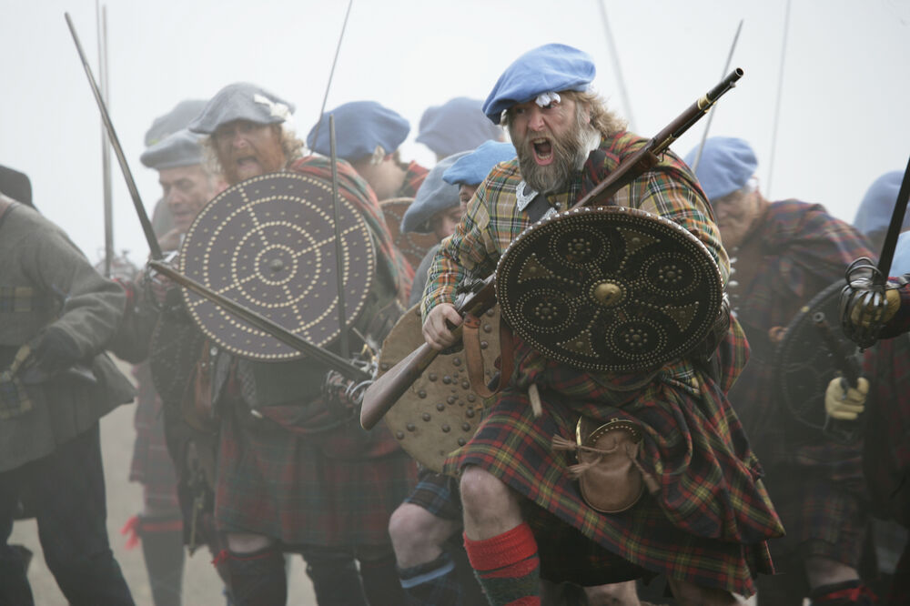 A group of men re-enact the Highland charge at the Battle of Culloden, dressed as Jacobite soldiers. They wear blue berets and carry large targes.