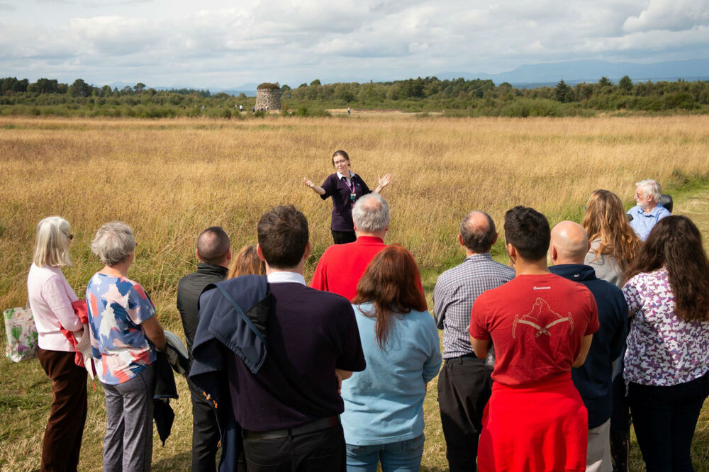 A woman faces a group of people all ages, giving them a tour of Culloden Battlefield. Behind her, the Culloden memorial cairn is visible.
