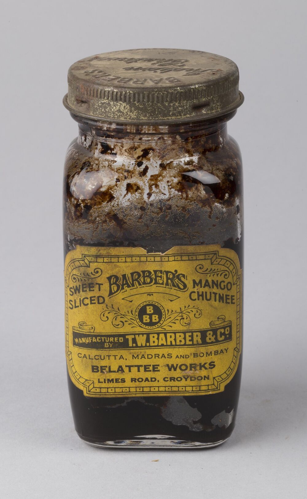 A very old glass jar, with a metal screw lid, is displayed against a plain grey background. Chutney can be seen smeared inside. A bright yellow label on the front says: Sweet sliced Barber's Mango chutnee. Manufactured by T W Barber & Co. Calcutta, Madras and Bombay. Belattee Works, Limes Road, Croydon.