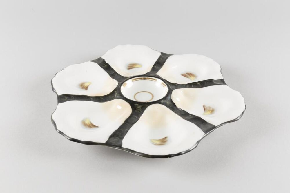 A dish used to serve oysters is displayed against a plain grey background. The dish almost resembles a flower, with six petals surrounding a round section in the middle.