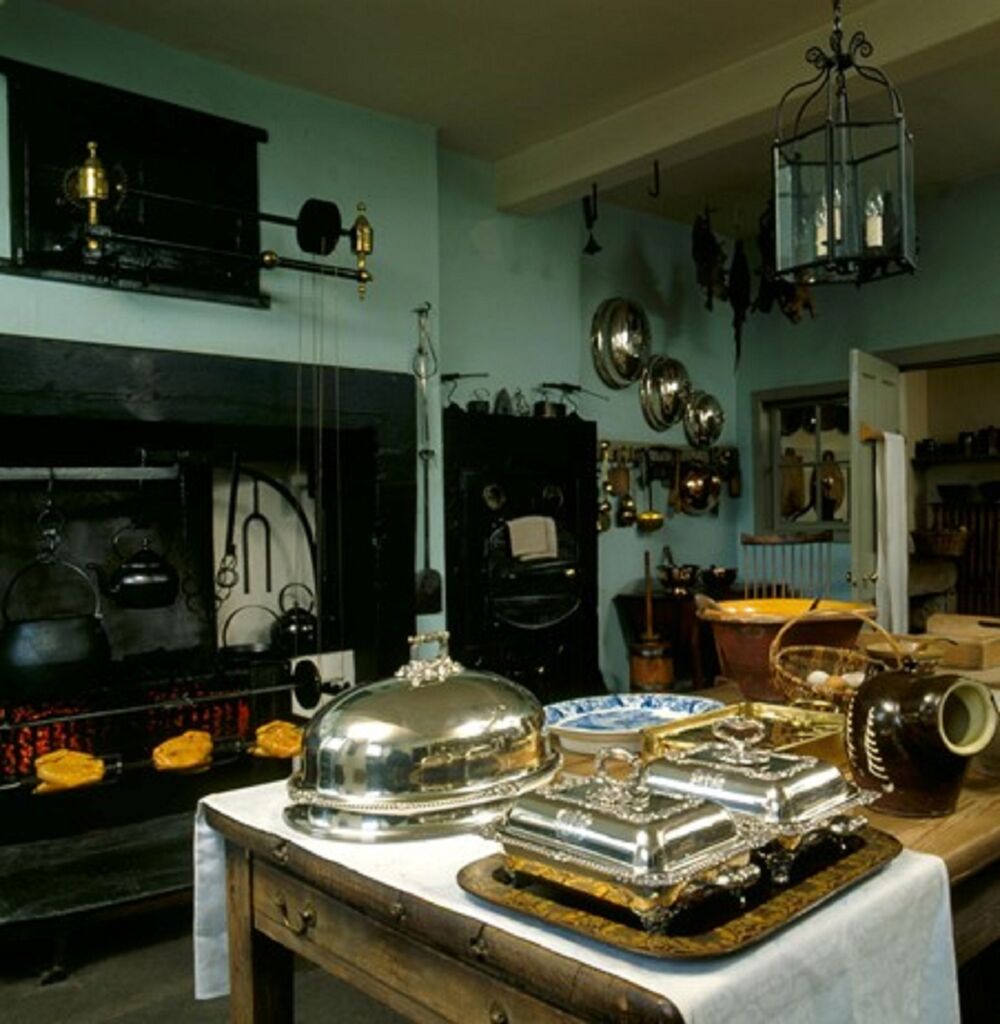 A view of the re-created kitchen at the Georgian House. A large table stands in the centre of the room, filled with platters, plates and pottery. Behind stands the range, with tin kettles and meat hanging over it. Other pots, pans and utensils hang from the walls.