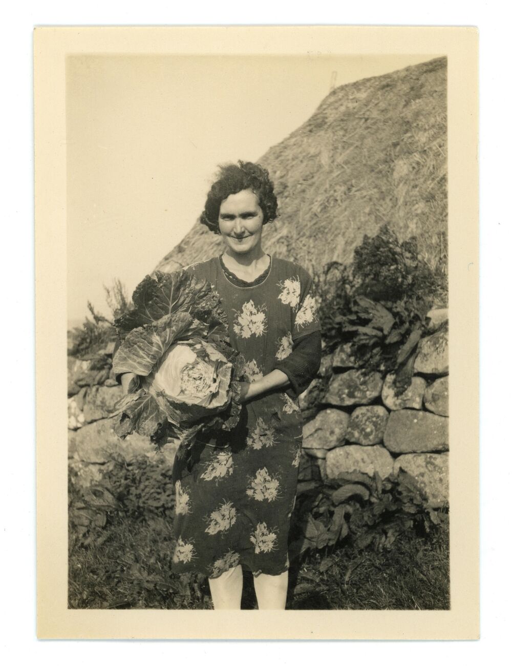 A black and white photo of a woman holding a very large cabbage, standing outside a thatched cottage. She is wearing a printed floral dress.