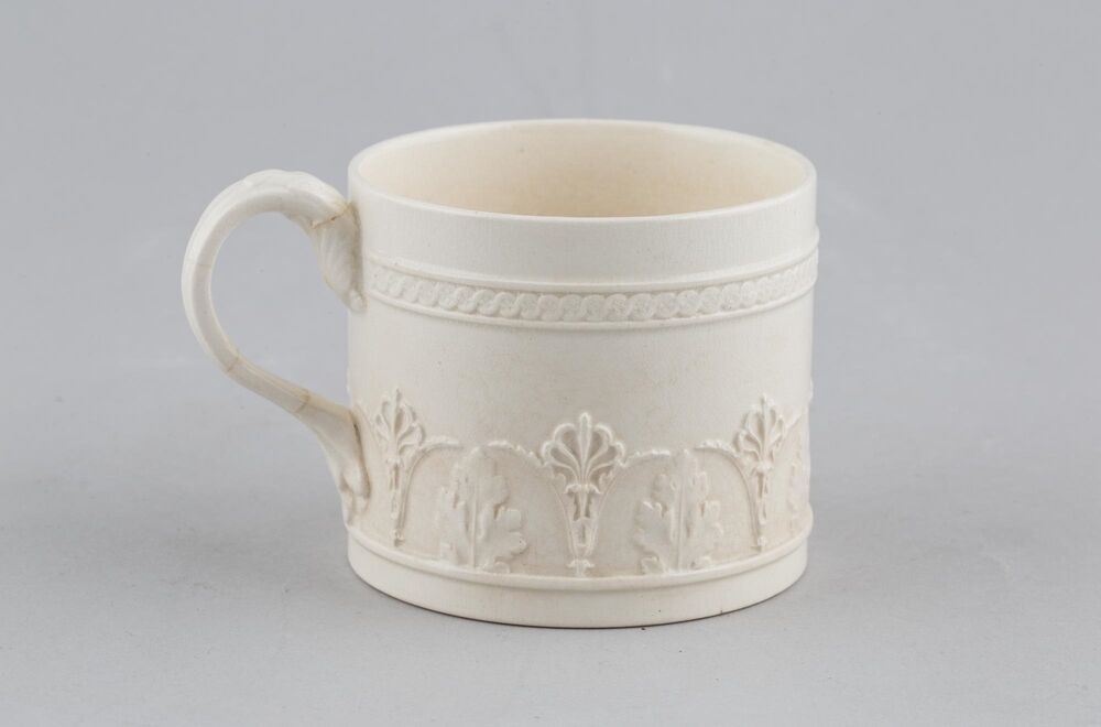 A small, white embossed coffee cup is displayed against a plain grey background. It is decorated with a band of palmettes and foliage and has a reeded handle.