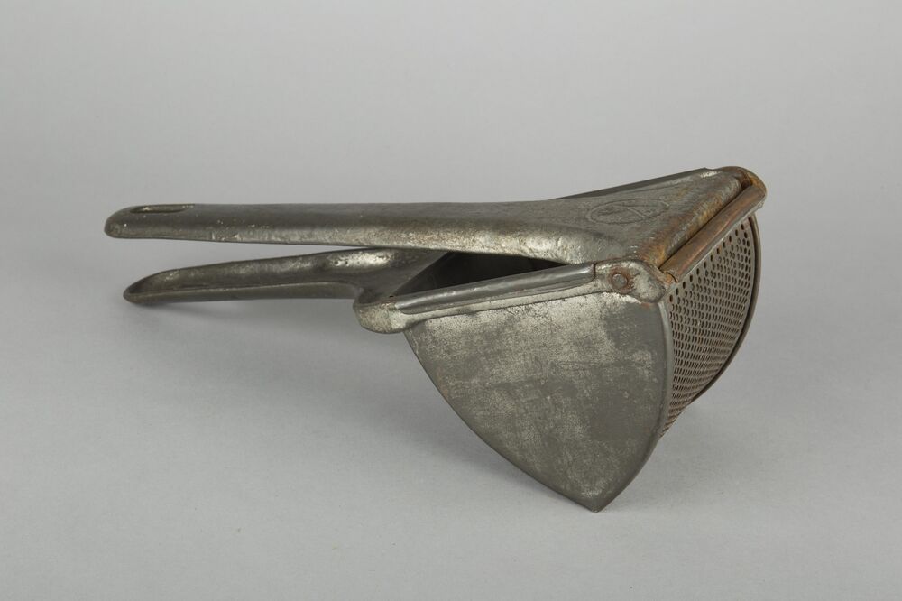 An old-fashioned metal vegetable masher, which looks a little like a garlic press, is displayed against a plain grey background.