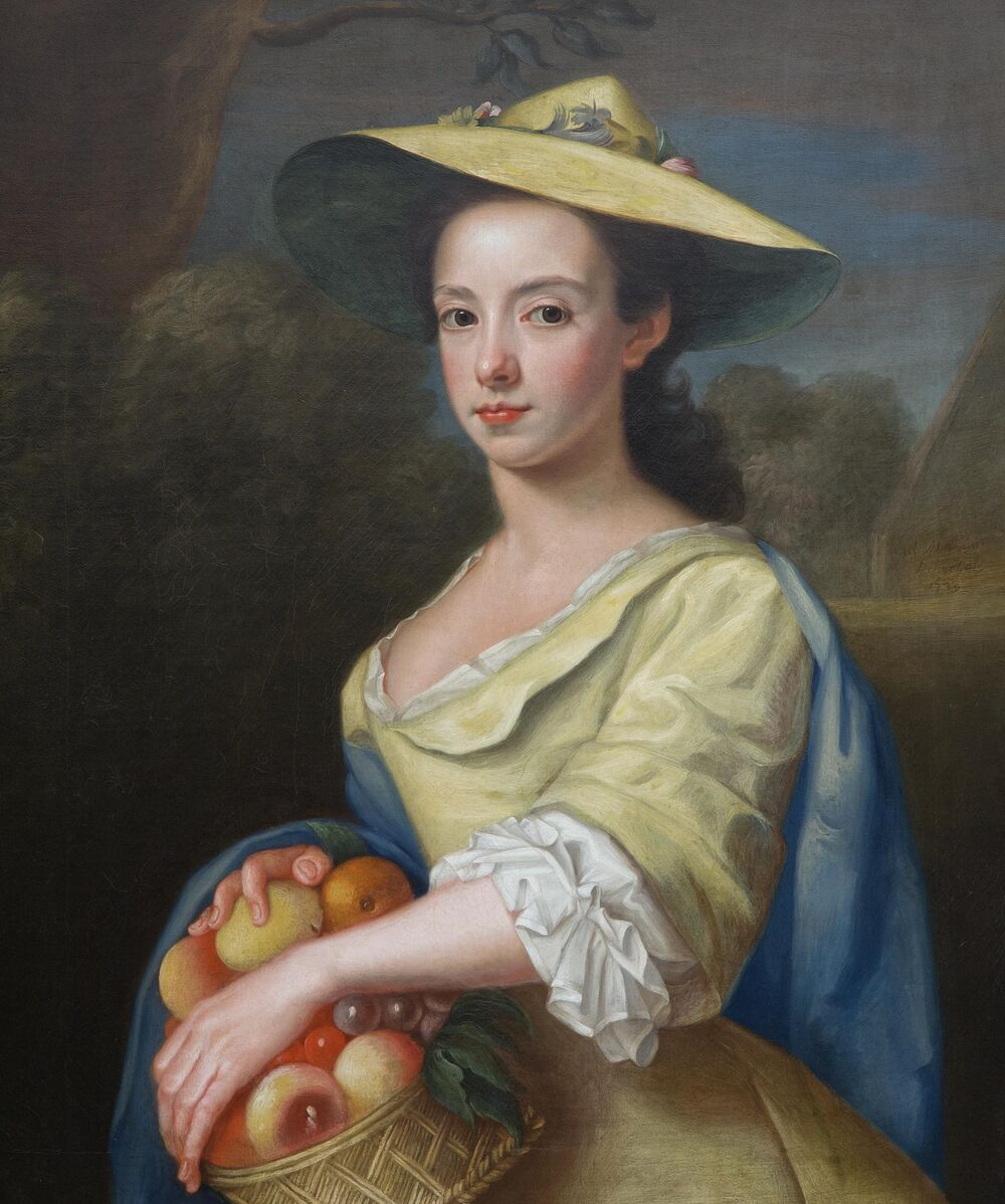 An oil portrait of a young Georgian woman holding a basket filled with peaches, grapes and citrus fruits. She wears a wide brimmed hat, a yellow dress and a blue shawl.