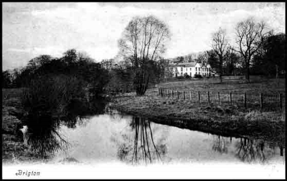 A black and white photograph of a large white house, seen almost in the distance. A still river is in the foreground, with wintery trees reflected in the water.