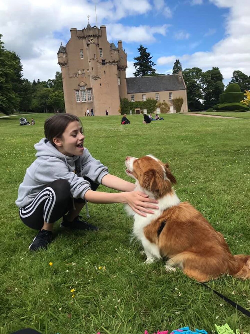 A smiling girl crouches down and pets a brown and white dog on the lawn in front of Crathes Castle. The dog appears to smile at the girl and sits nicely, her lead lying on the grass beside her.