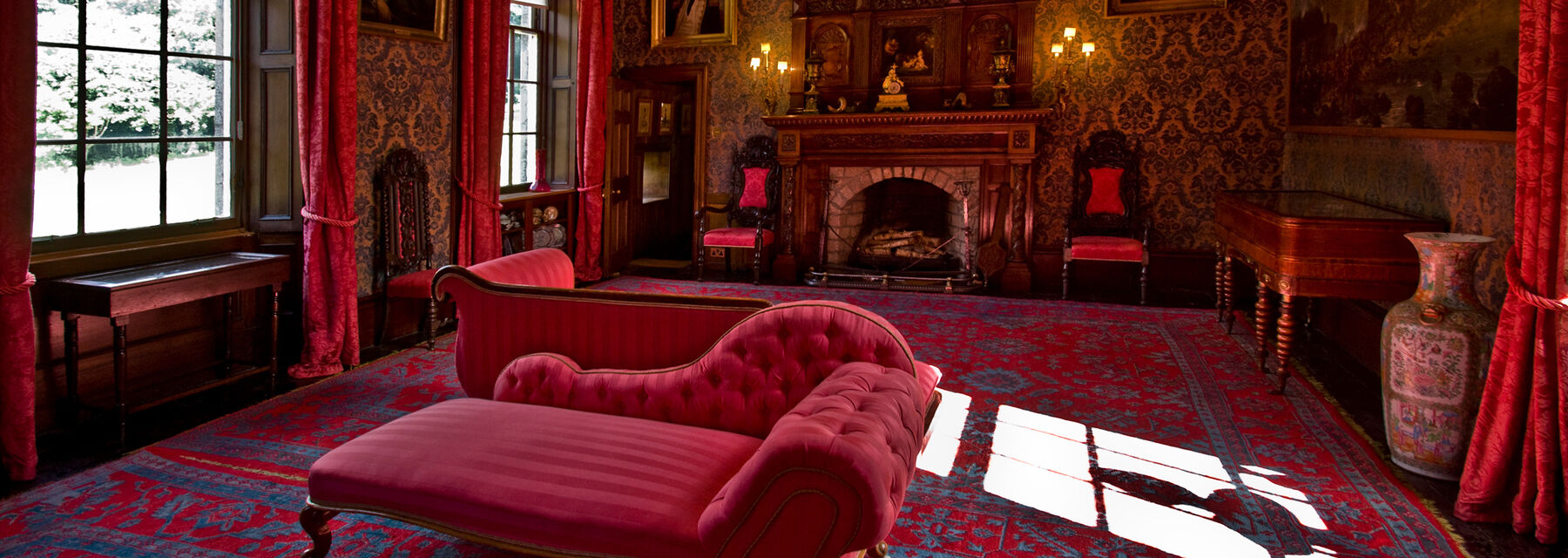 A view of a grand drawing room, with many red furnishings. Oil paintings hang on the patterned walls.