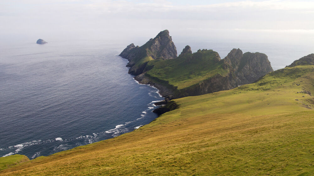 A view of the island of Hirta, part of St Kilda, looking out to sea. In the foreground is a steep grassy cliff. It stretches to a rocky point, reaching out into the sea.