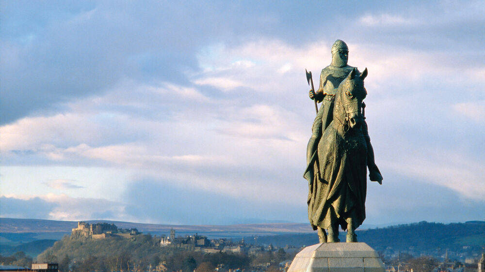 A statue of Robert the Bruce on horseback faces the camera, with the Stirling hills and blue skies in the background.