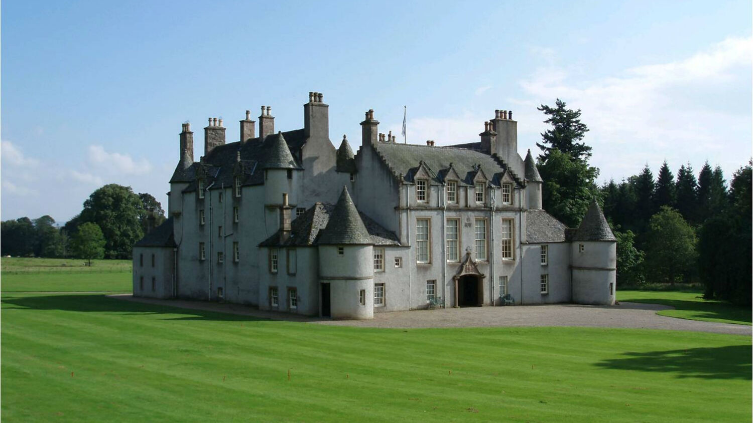 A view of the exterior of Leith Hall, from across some manicured lawns. It is a large stately home, with grey walls and a slate roof, with little towers and turrets at each corner.
