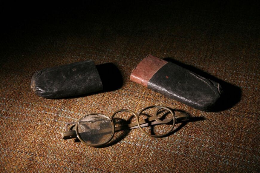 A very old pair of glasses, with one glass lens missing, lies on an old wooden table. An old leather case is in two halves behind them. The lighting is quite low in the photograph.