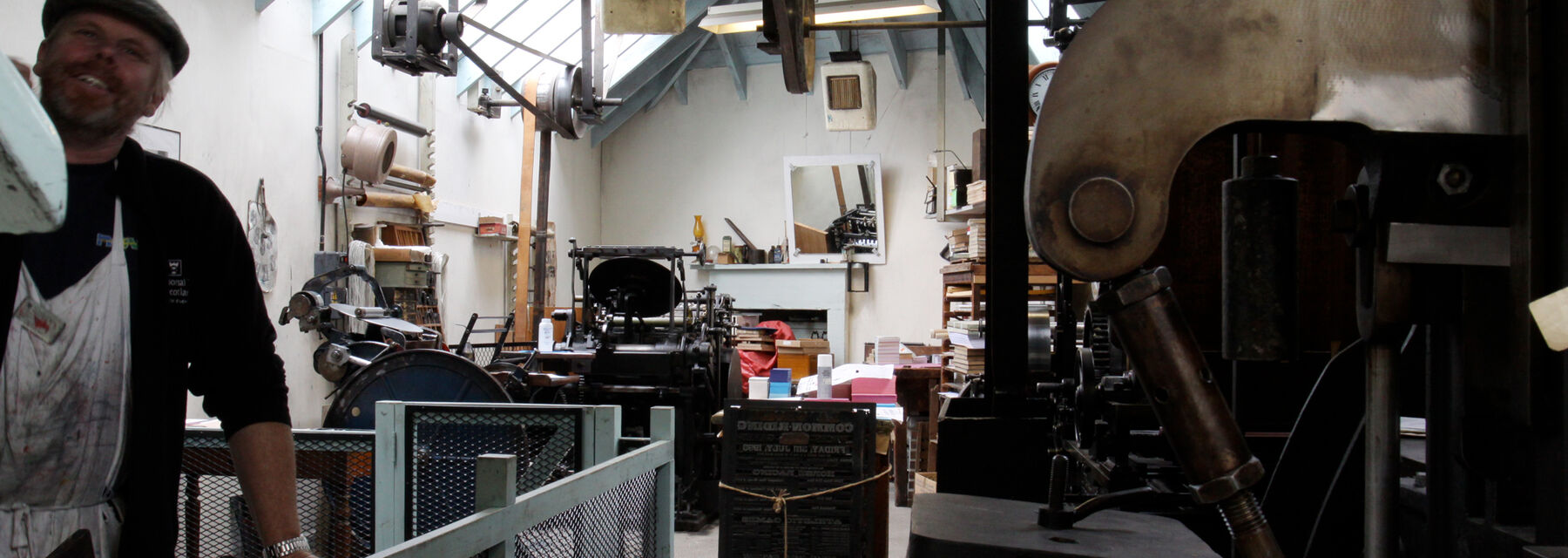 The workshop at Robert Smail's. A beamed roof lets in light, and the workshop is crammed with letterpress machinery and tools, some hanging from above. A man leans on a rail.