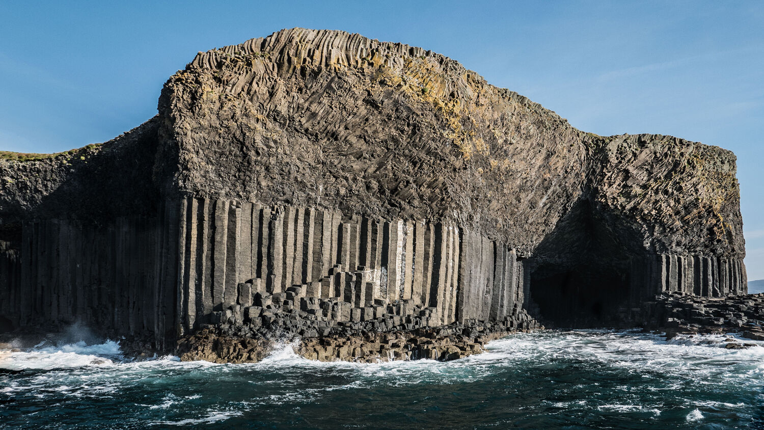 A view of a deep cave in a cliff, seen from the sea on a sunny day. The cliffs have remarkable basalt columns.