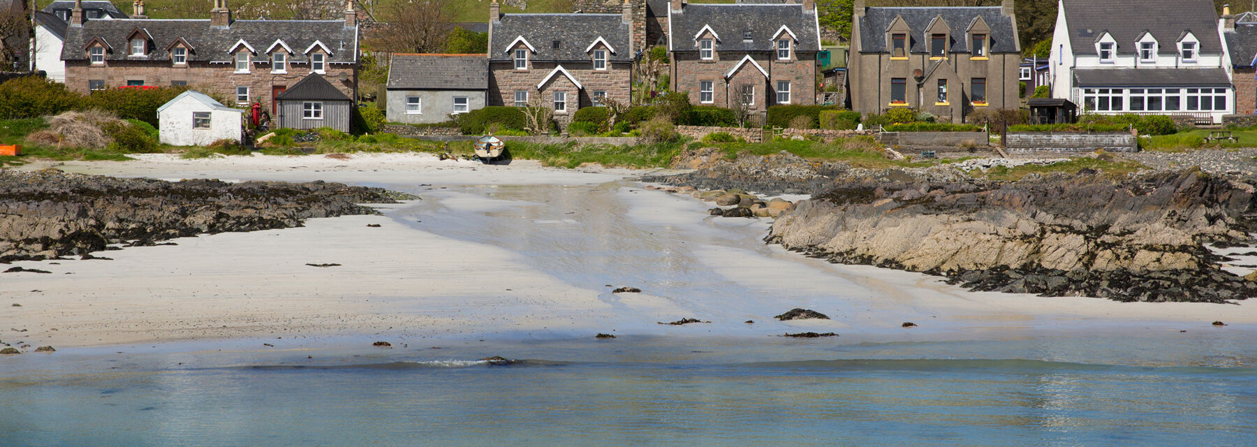 A white sandy beach on Iona, with the turquoise sea in the foreground. Houses and a hill are in the background.